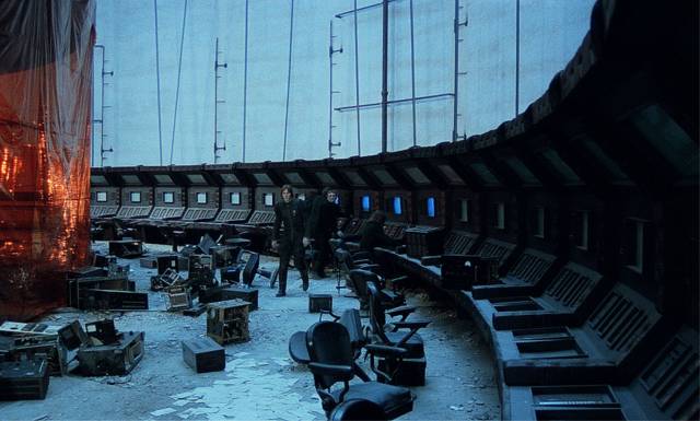 A large room with old computer terminals lining the circular perimeter. The floor is covered in debris and partially-disassembled computers. Three people dressed in black investigate the scene.